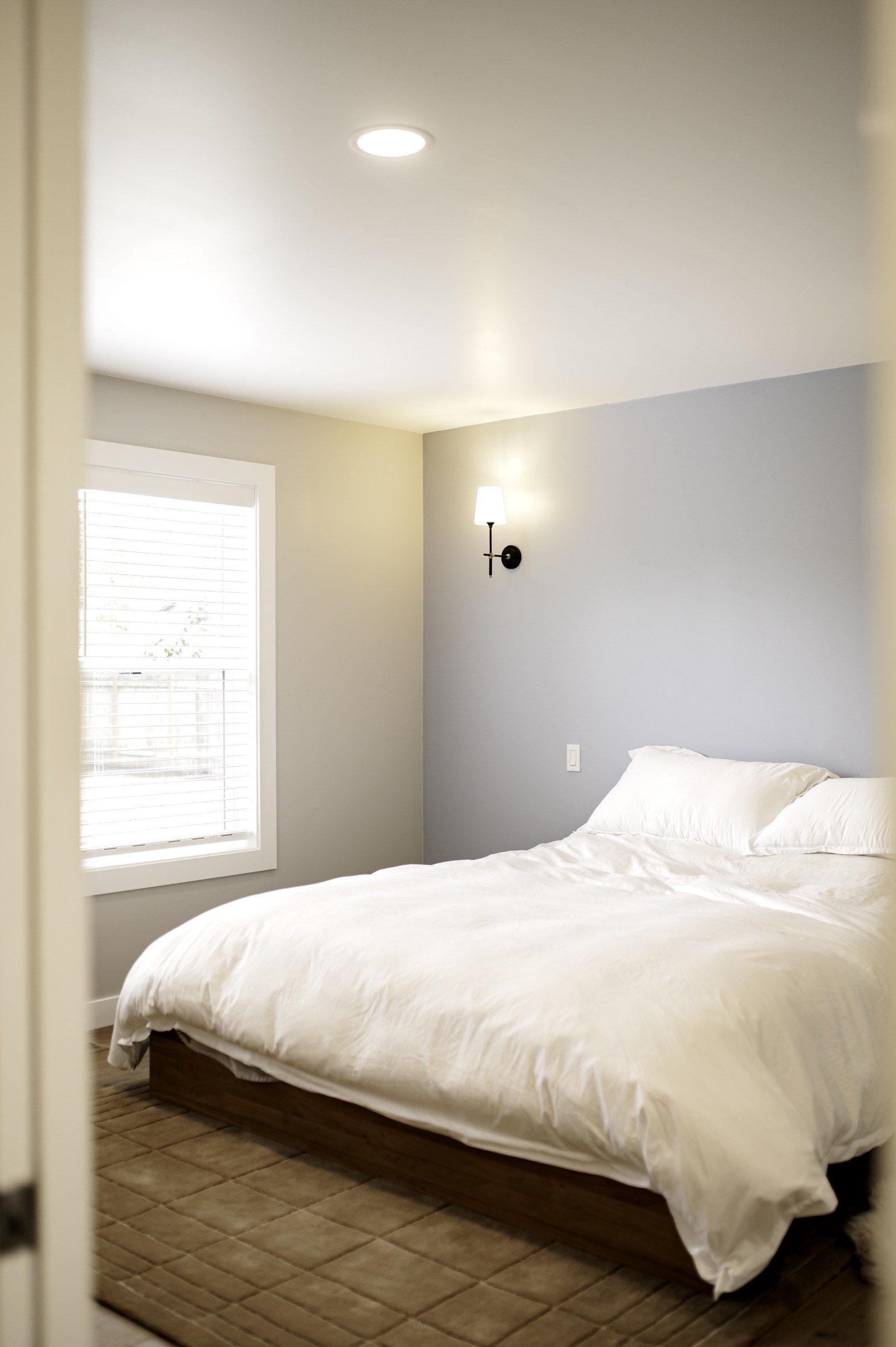 Finished bedroom with white bedding, a gray accent wall, window trim and a wall sconce