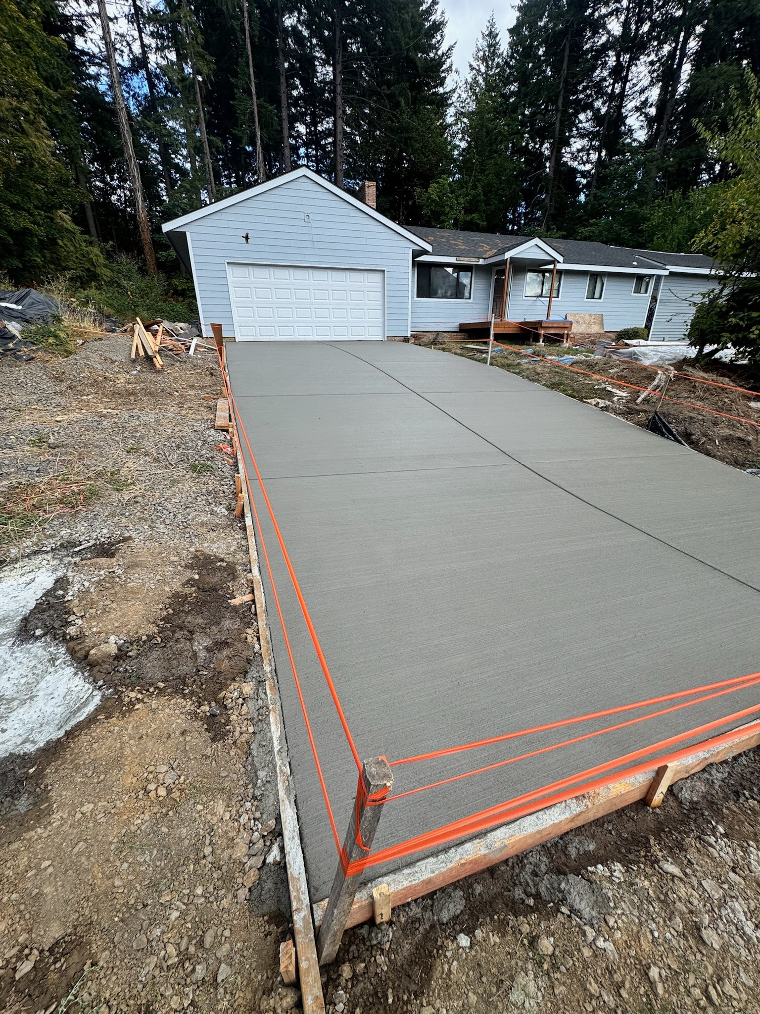 Newly placed concrete driveway leading to a garage, with temporary formwork and unfinished ground along the sides.