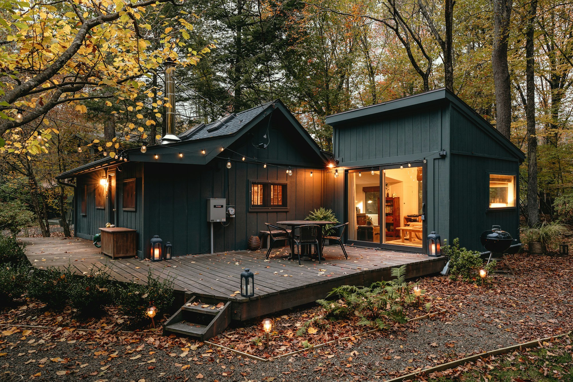 Dark timber cabin with a dining deck and warm interior lighting beside autumn trees