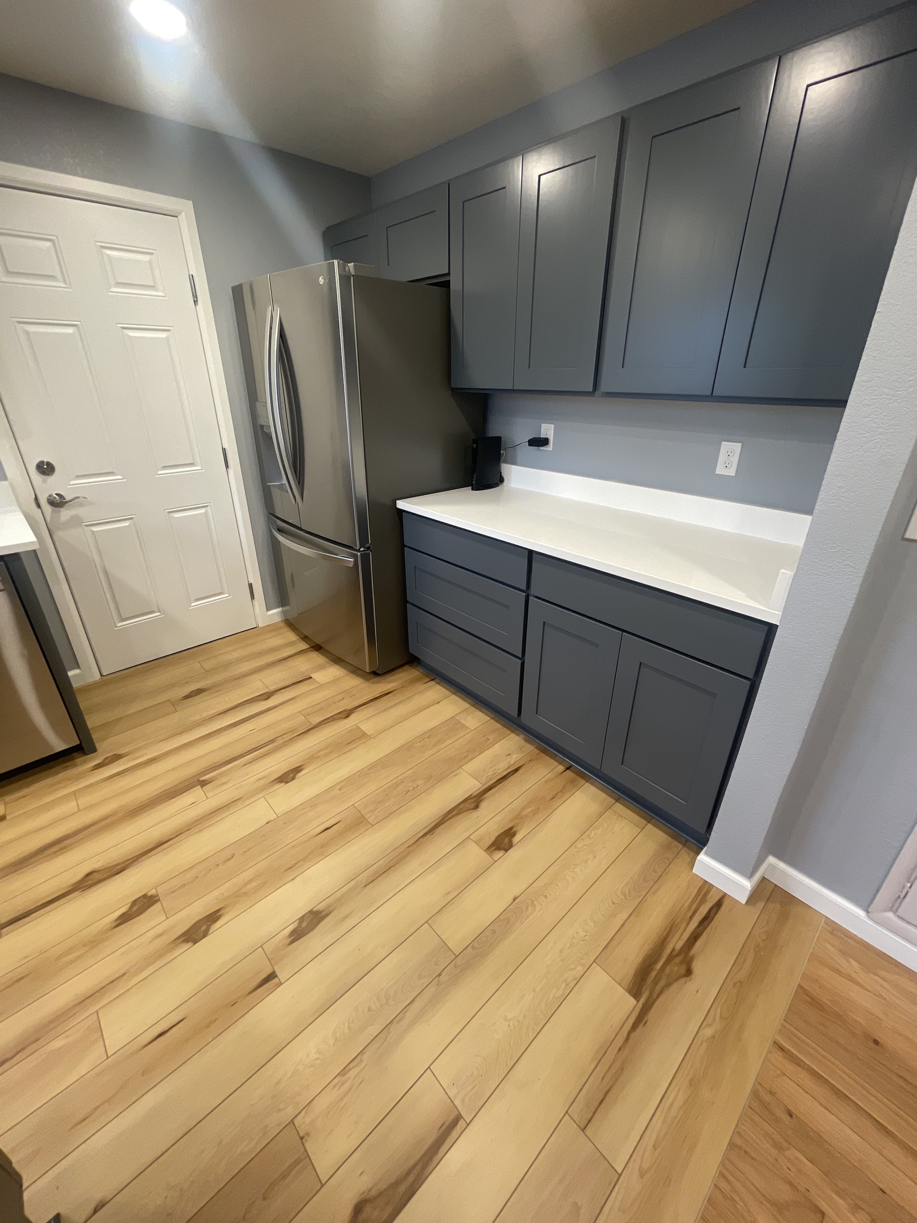 Gray kitchen storage cabinets beside a refrigerator, white door, and wood flooring