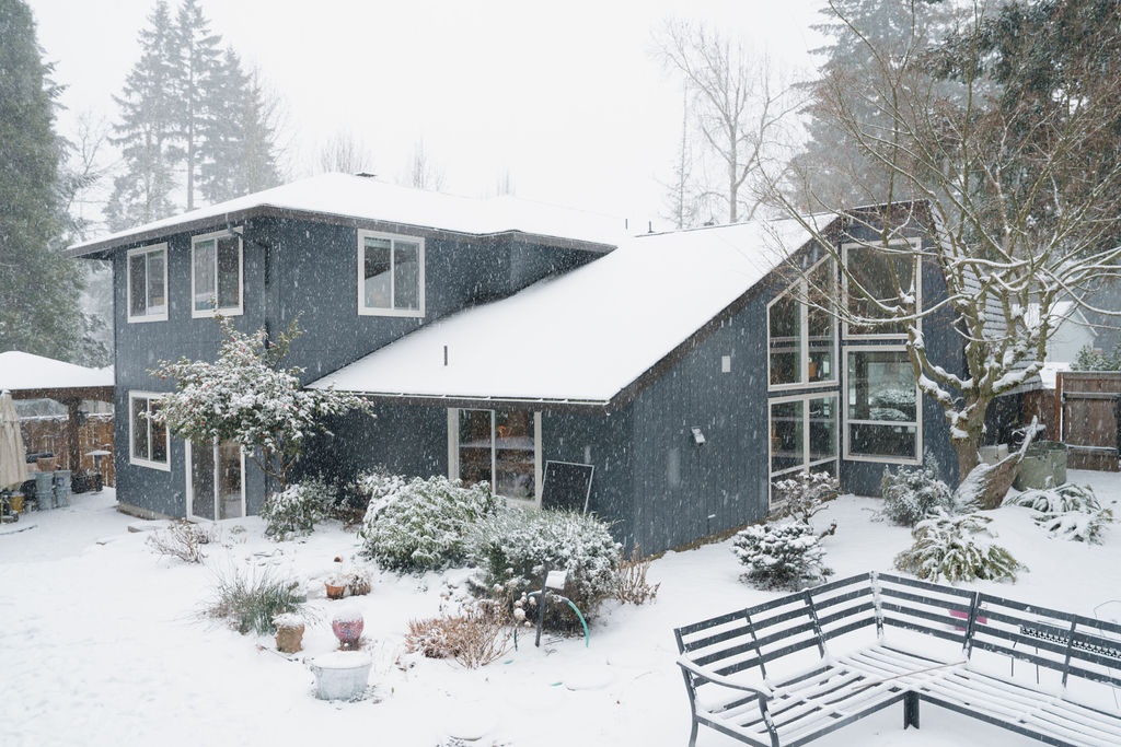 Rear elevation with blue siding, a two-story volume and a lower glazed room beneath snow-covered roofs